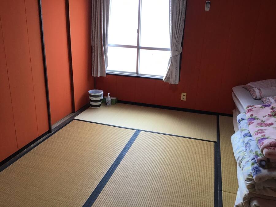 Een kamer met tatami vloerbedekking, een bed met bloemenprint beddengoed, een raam met gordijnen, en een kleine prullenbak.