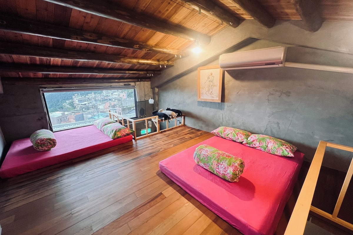 A cozy room with wooden floors, two beds with floral bedding, a window with a view of buildings, and a mounted air conditioner.