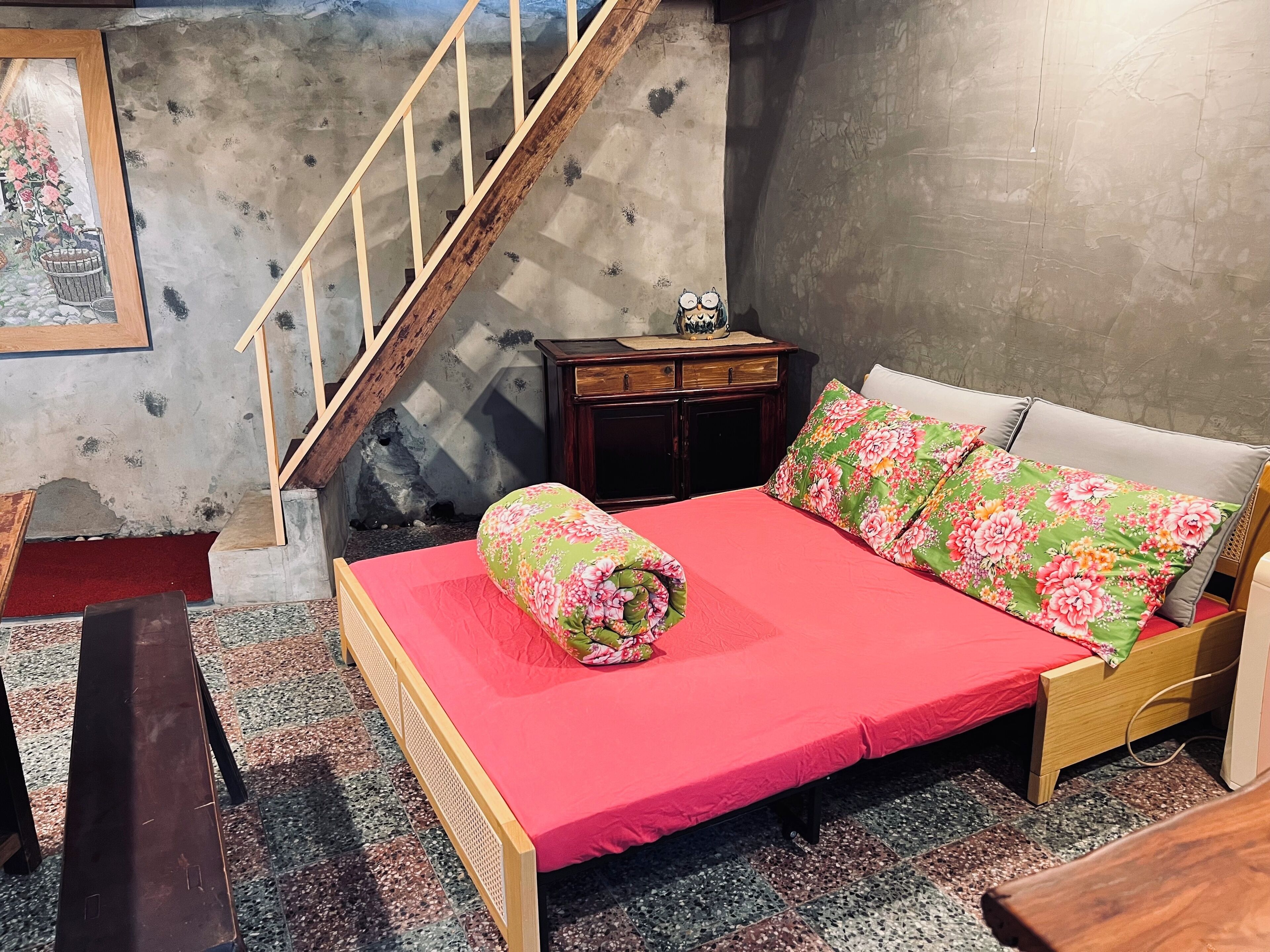A bedroom with a wooden bed, a red bedspread, floral pillows, a wooden nightstand, a dresser, and a staircase.