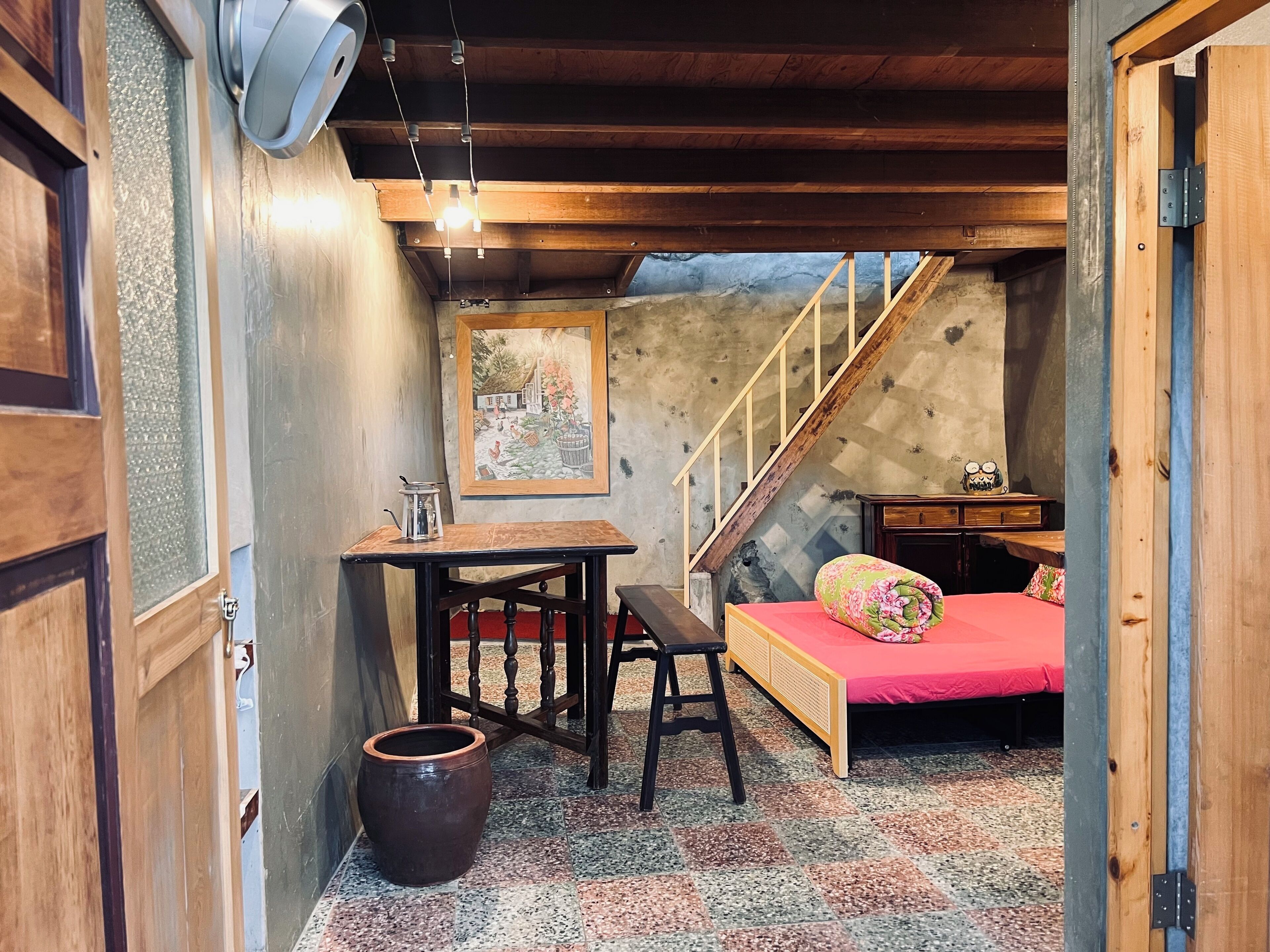 A cozy room with a wooden ceiling, a bed with a red bedspread, a small table with a kettle, a chair, and a large pot on the floor.