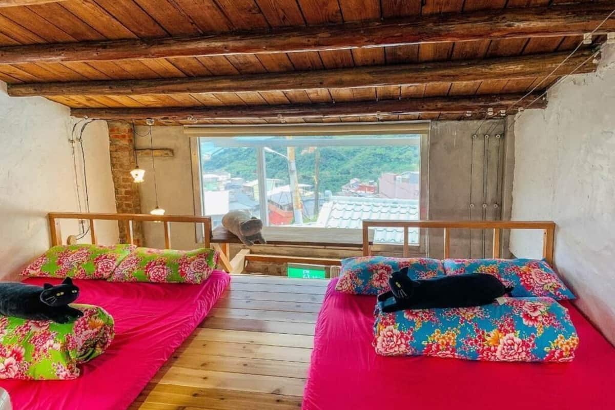 A room with two beds, wooden flooring, a sloped ceiling, and a window with a view of buildings.