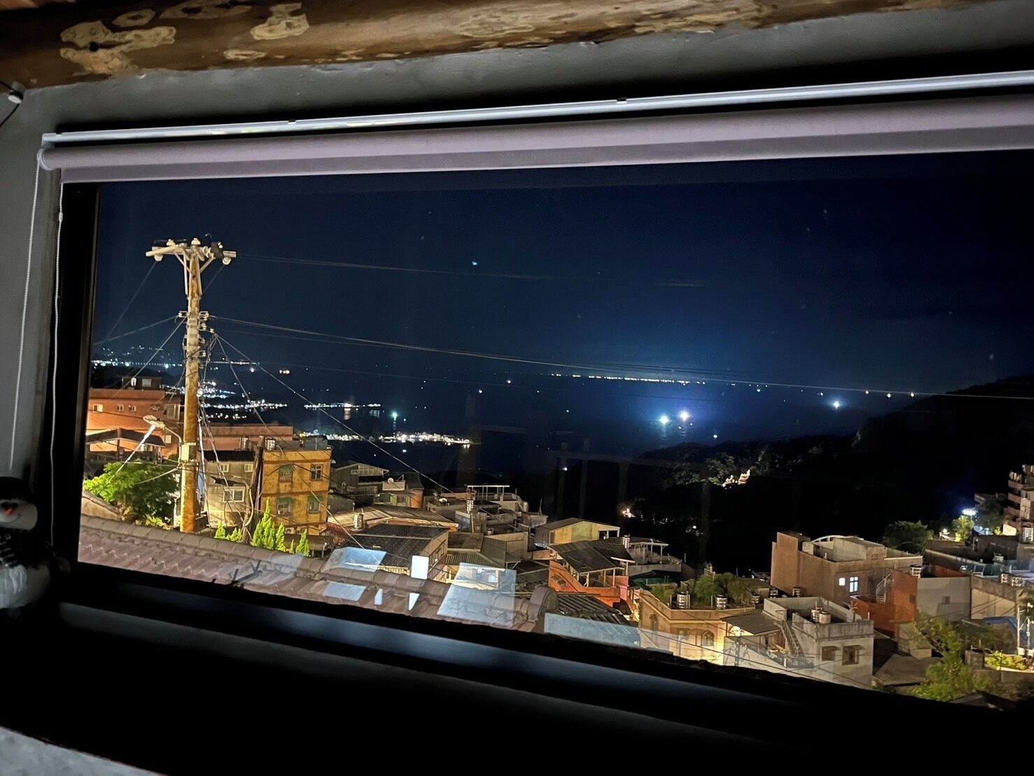 A nighttime cityscape viewed through a window, featuring illuminated buildings, a prominent utility pole, and a distant bridge.