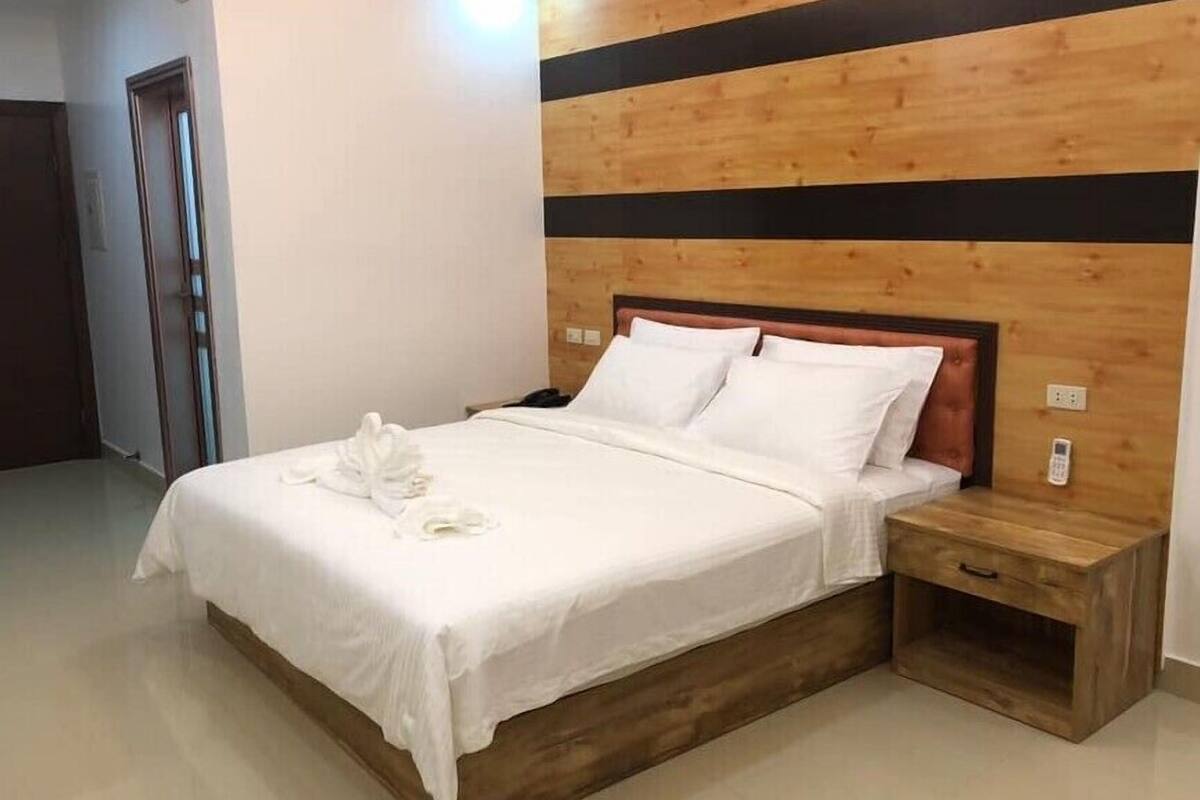 Ein ordentlich bezogenes Bett mit weißen Leinen und einem Handtuch, ein Holz-Kopfteil mit schwarzem Streifen sowie ein Nachttisch mit Schublade.