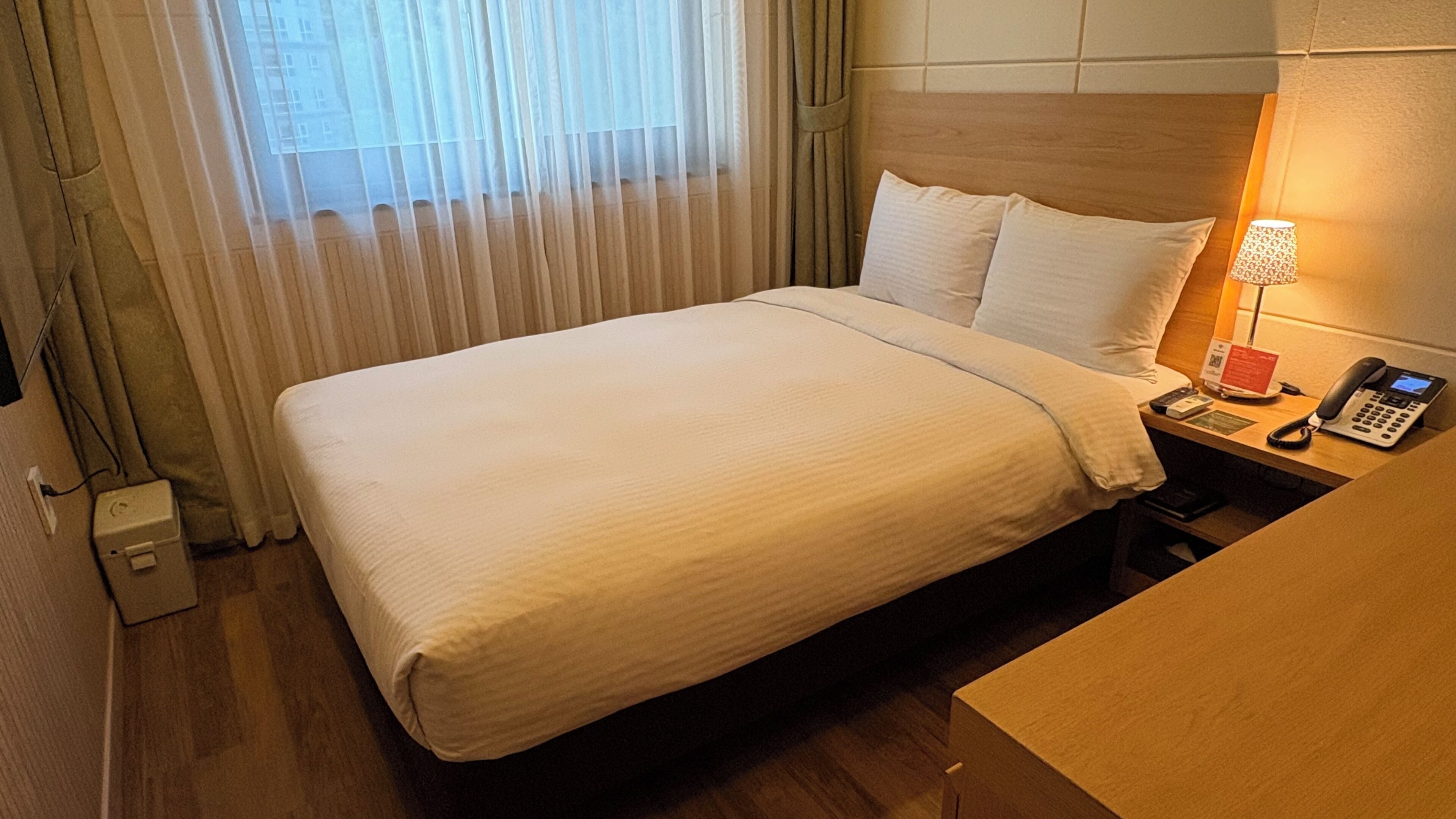 A hotel room with a bed, a bedside table with a lamp, a phone, and a small box.