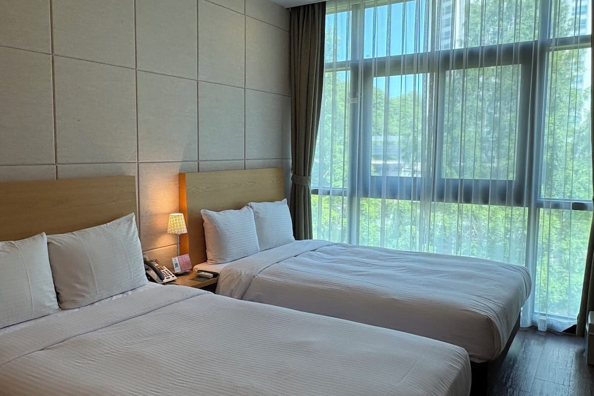 Habitación de hotel con dos camas, cabecero, mesita de noche con lámpara y ventana con cortinas.
