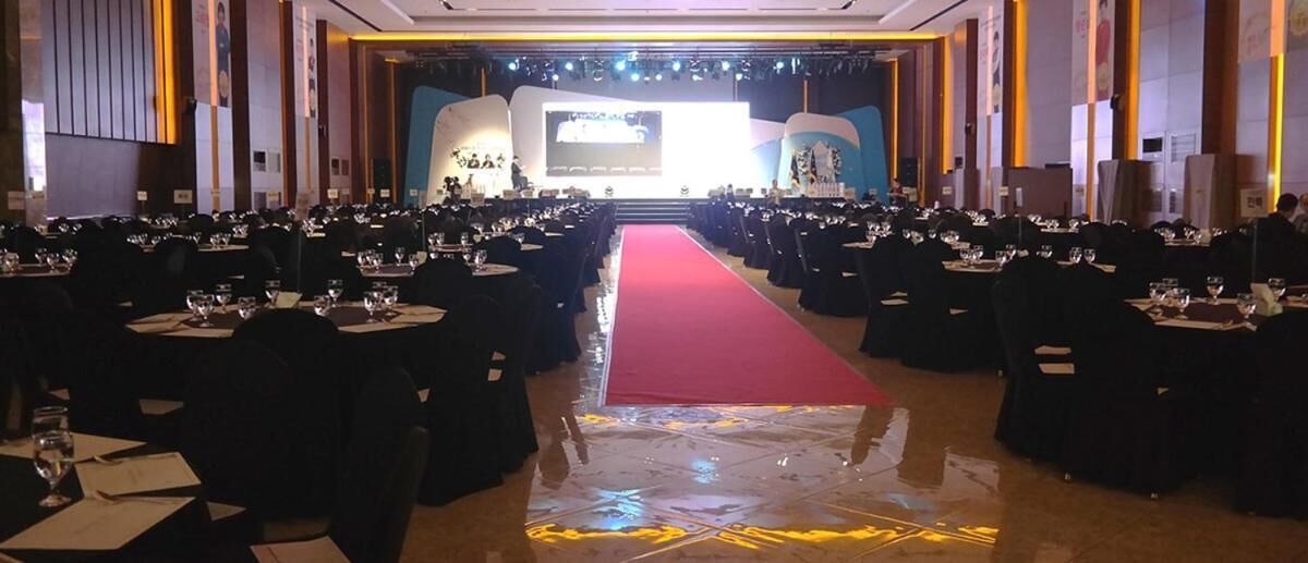 Un salón de banquetes con mesas dispuestas en filas, una alfombra roja y un escenario al frente.