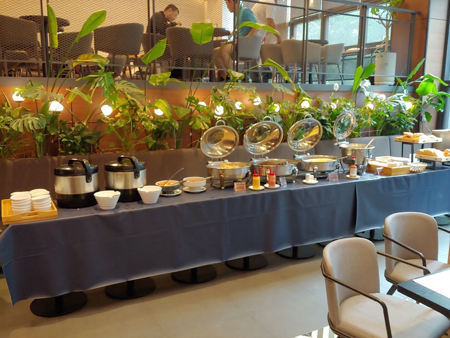 A buffet setup with various food items and condiments, surrounded by potted plants and seating areas.