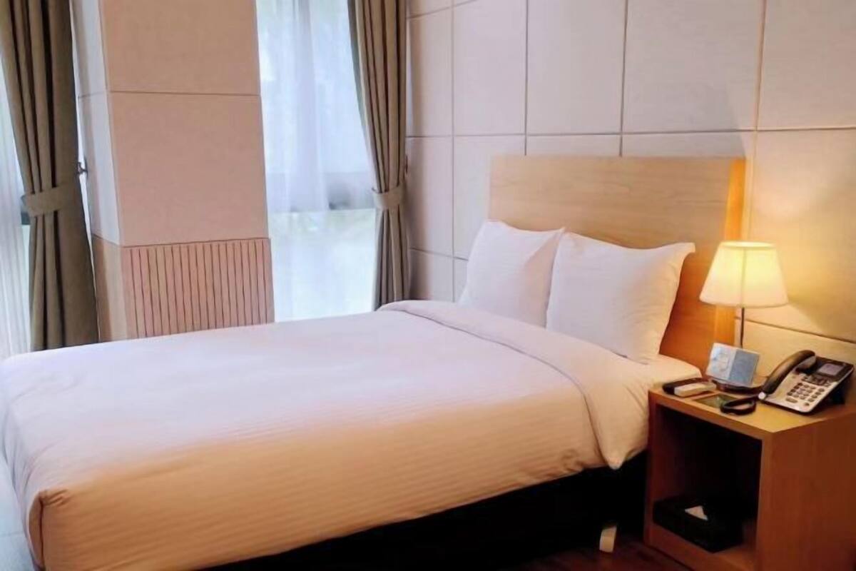 Habitación de hotel con cama, mesita de noche con lámpara, teléfono y ventana con cortinas.
