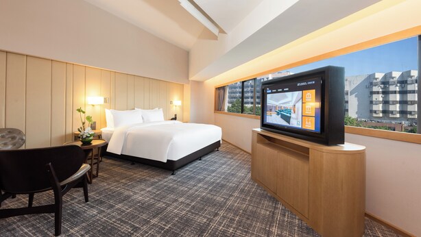 A hotel room with a bed, a TV on a wooden stand, a desk with a lamp, and a view of buildings through a window. - Xence Hotel (Xiamen)