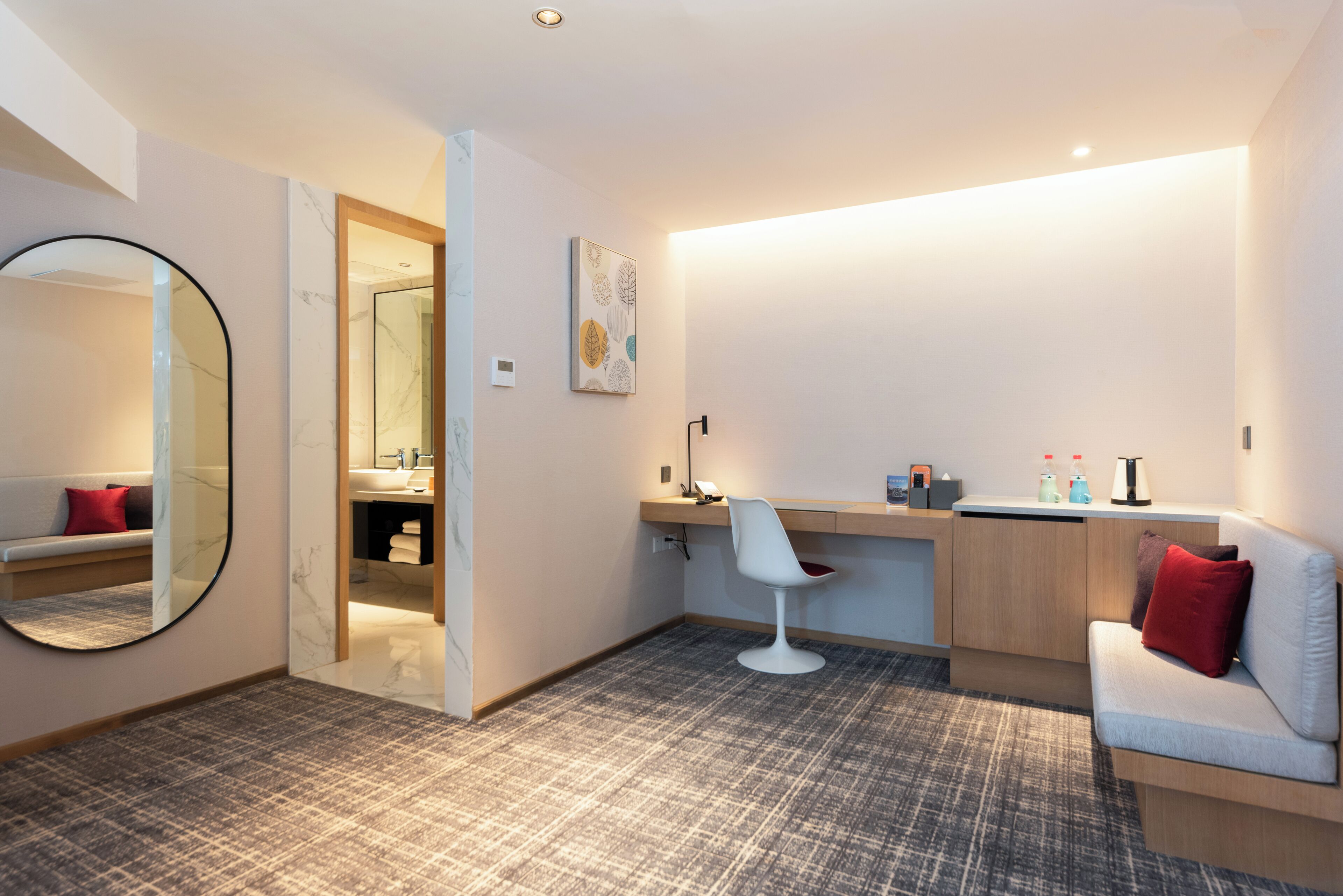 A modern hotel room with a sofa, a desk, a chair, a mirror, and a bathroom visible through an open door.