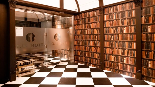 A hotel lobby with a checkered floor, a glass elevator, and a wall of bookshelves. - T.O Hotel Main Station (Taipei)