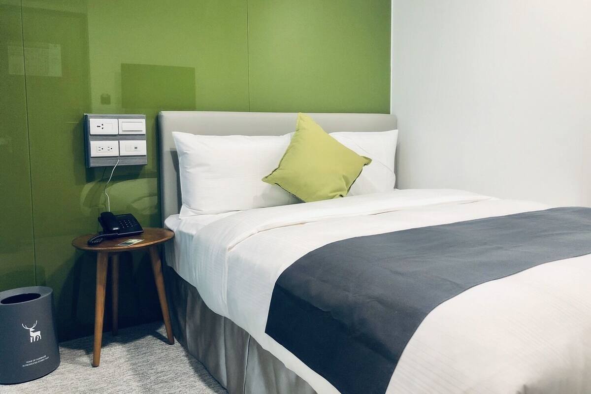 A hotel room with a bed, a bedside table with a phone, and a green wall.