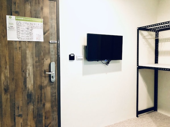 Una habitación con puerta de madera, televisor montado, estantería metálica y pared blanca.