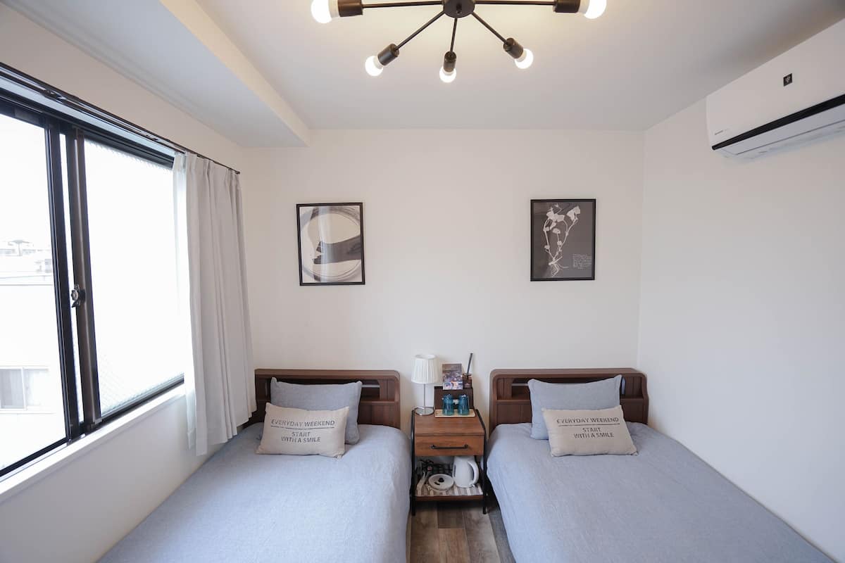 A hotel room with two single beds, a nightstand between them, a ceiling light fixture, and two wall-mounted air conditioning units.