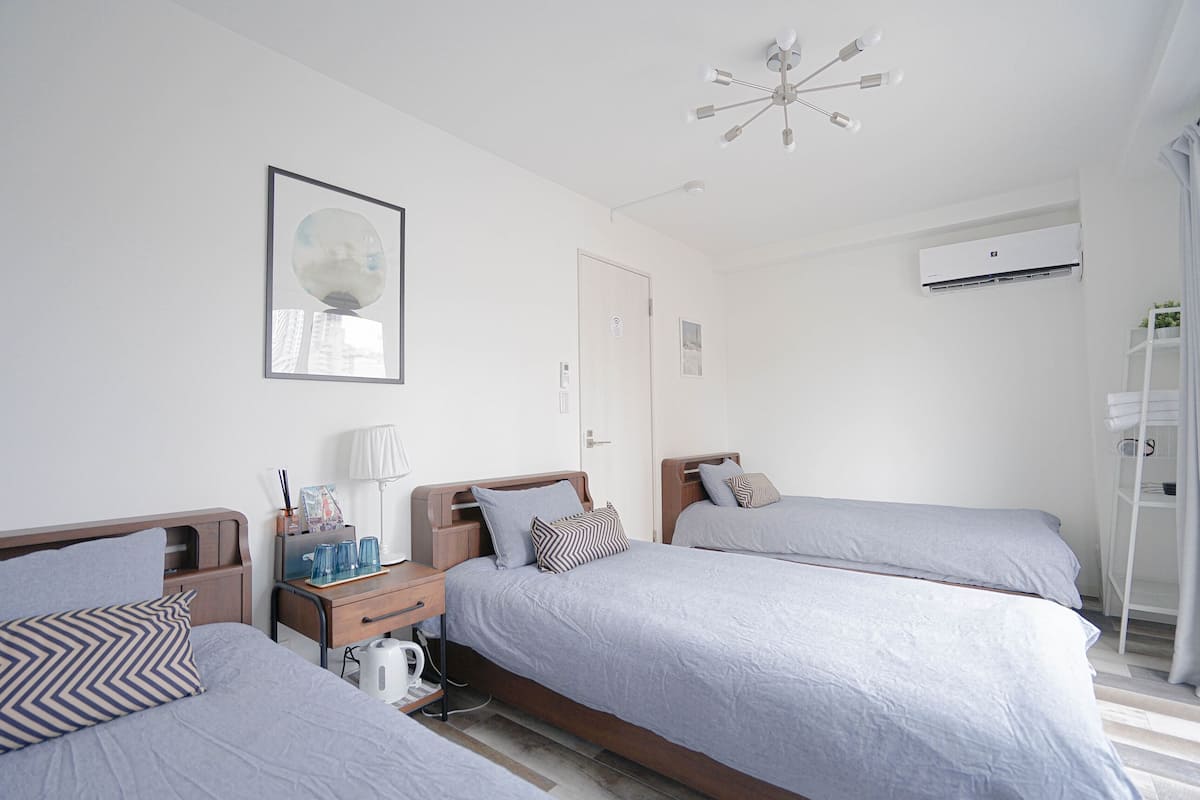 A modern hotel room with three single beds, a bedside table with a lamp, a coffee maker, and a framed picture on the wall.