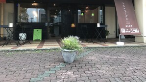 Entrada de restaurante com toldo preto, mesas ao ar livre e um vaso de planta em primeiro plano.