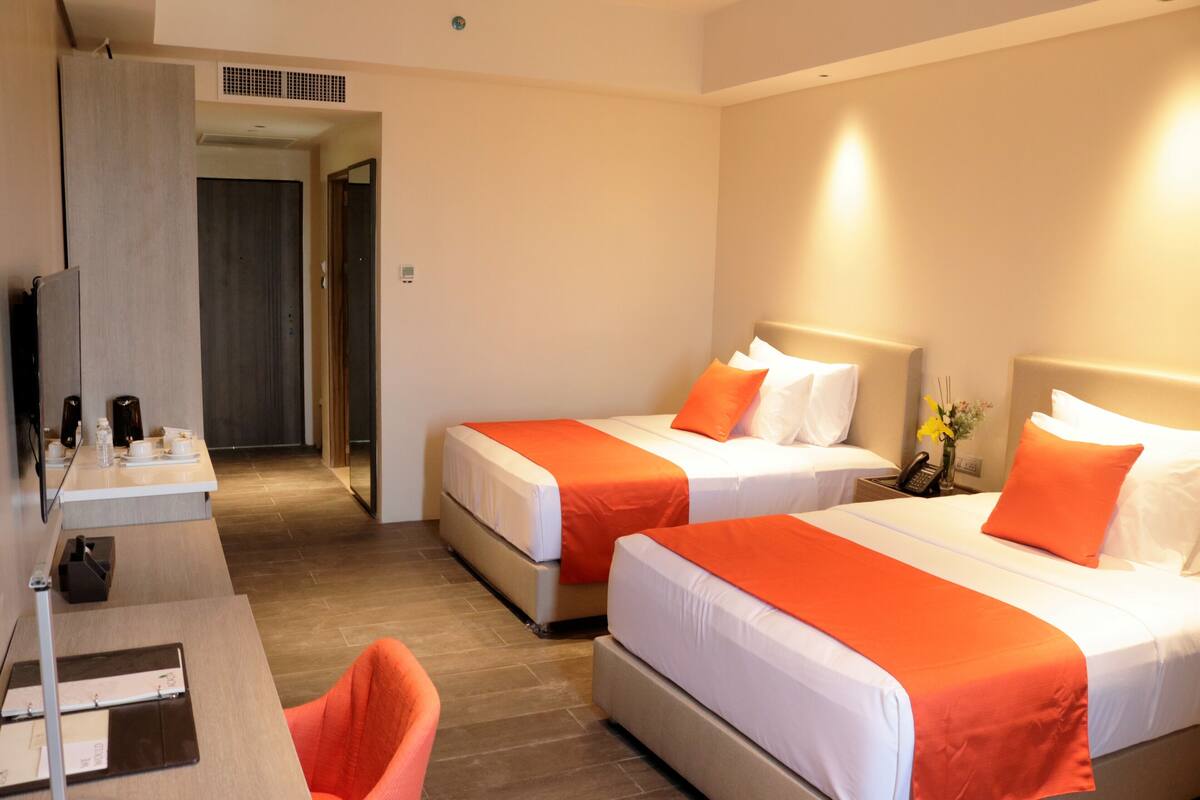A hotel room with two beds, each with white bedding and orange accents, a desk with a chair, and a small table with a phone and a vase of flowers.