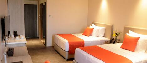 Habitación de hotel con dos camas, cada una con ropa de cama blanca y detalles en naranja, un escritorio con una silla y una mesita con un teléfono y un florero.