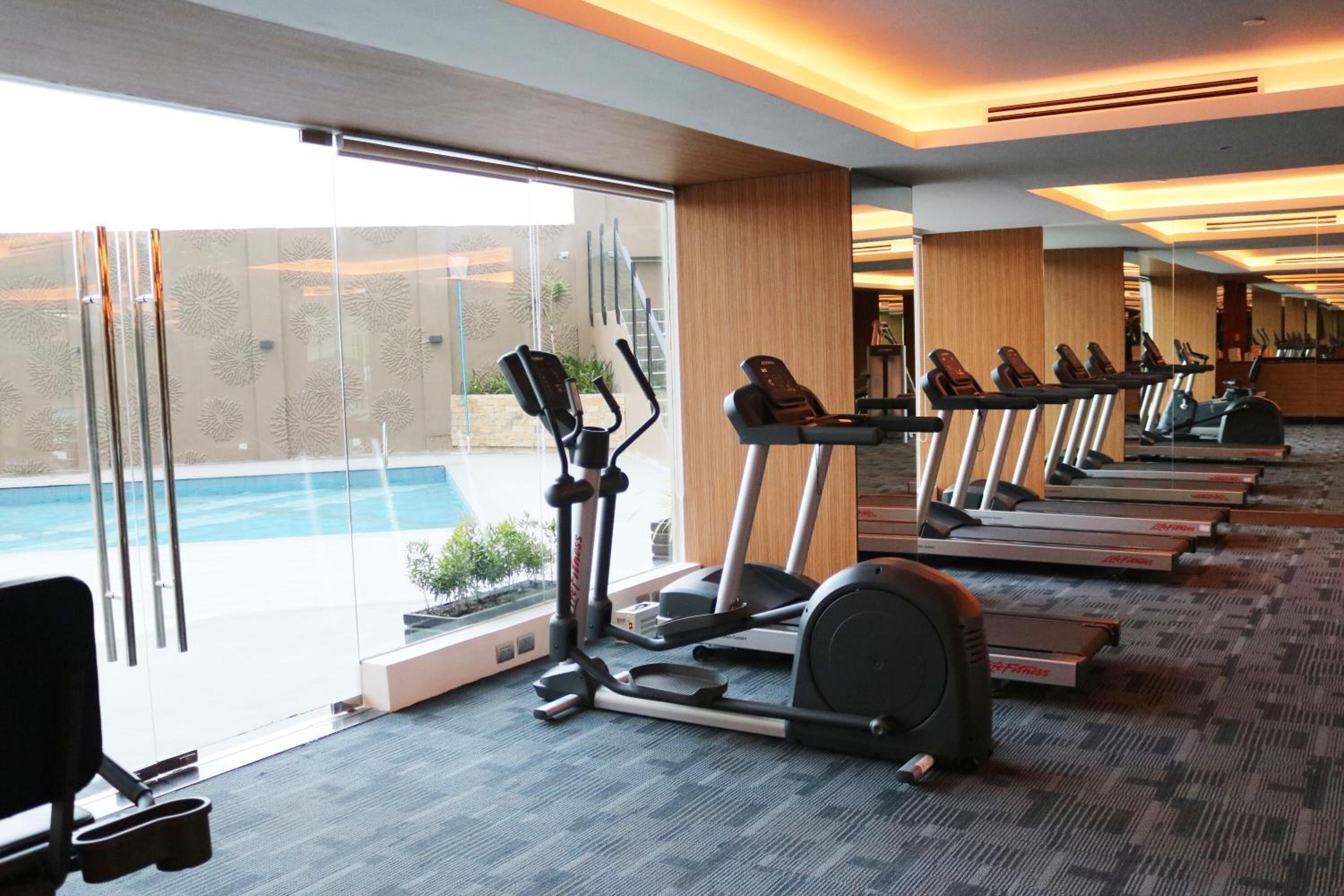 Un gimnasio con cintas de correr y una máquina elíptica, amplias ventanas y vistas a una piscina.