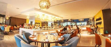 Un restaurant moderne avec des tables rondes et des chaises bleues, un coin bar et une cuisine ouverte visible au fond de la salle.