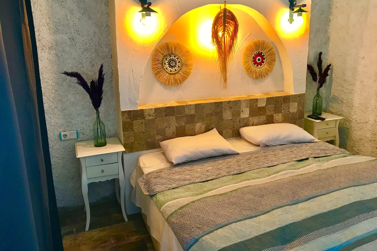 A bedroom with a bed, two nightstands, a wall-mounted light fixture, and decorative wall art.