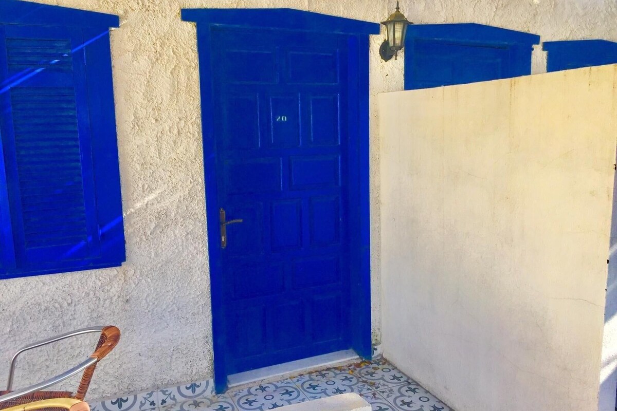 Um edifício com porta e persianas azuis, parede branca e chão de azulejos estampados.