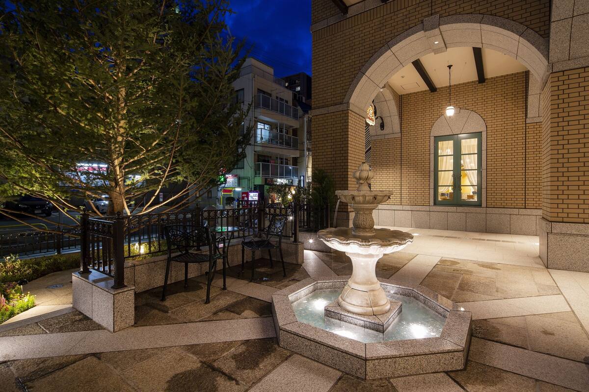 An outdoor patio area with a stone fountain, seating, and a building with a brick facade and arched entrance.