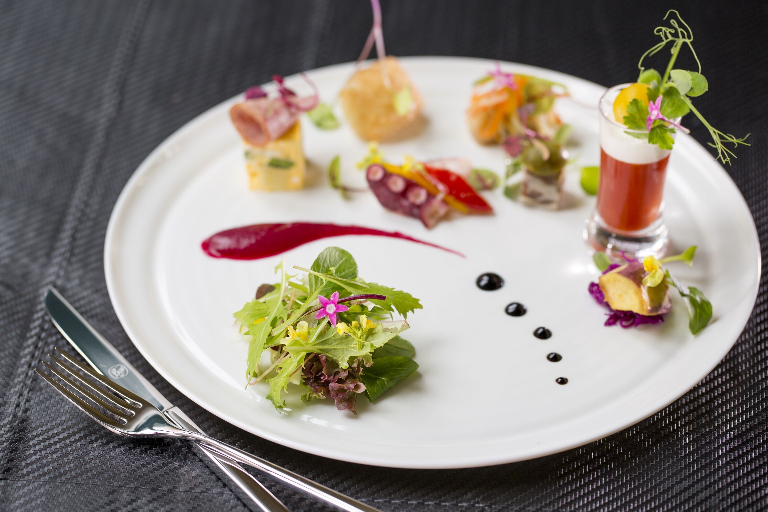 A white plate with various food items and a glass of red beverage, accompanied by a fork and knife on a dark textured surface.