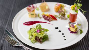 A white plate with various food items and a glass of red beverage, accompanied by a fork and knife on a dark textured surface.