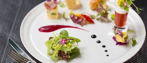 A white plate with various food items and a glass of red beverage, accompanied by a fork and knife on a dark textured surface.
