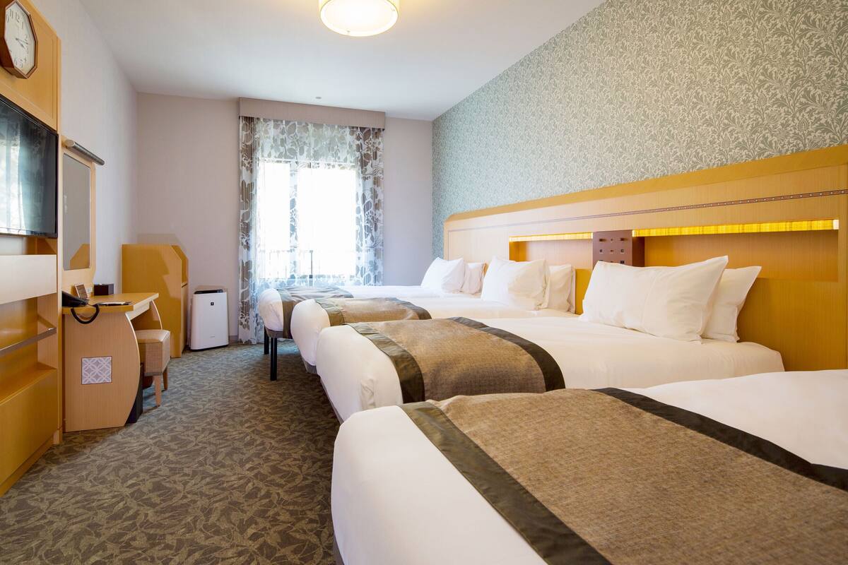 A hotel room with three beds, a desk, a chair, a clock, and a window with curtains.