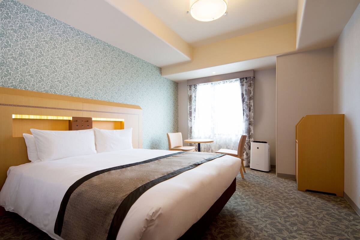 A hotel room with a large bed, a small table, a chair, and a wardrobe.