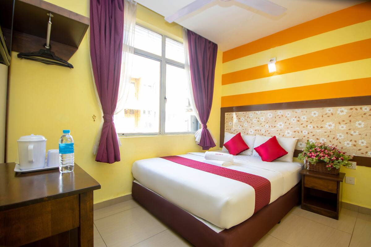 Quarto de hotel com cama, mesa de cabeceira, escrivaninha, garrafa de água, cafeteira e janela com cortinas.