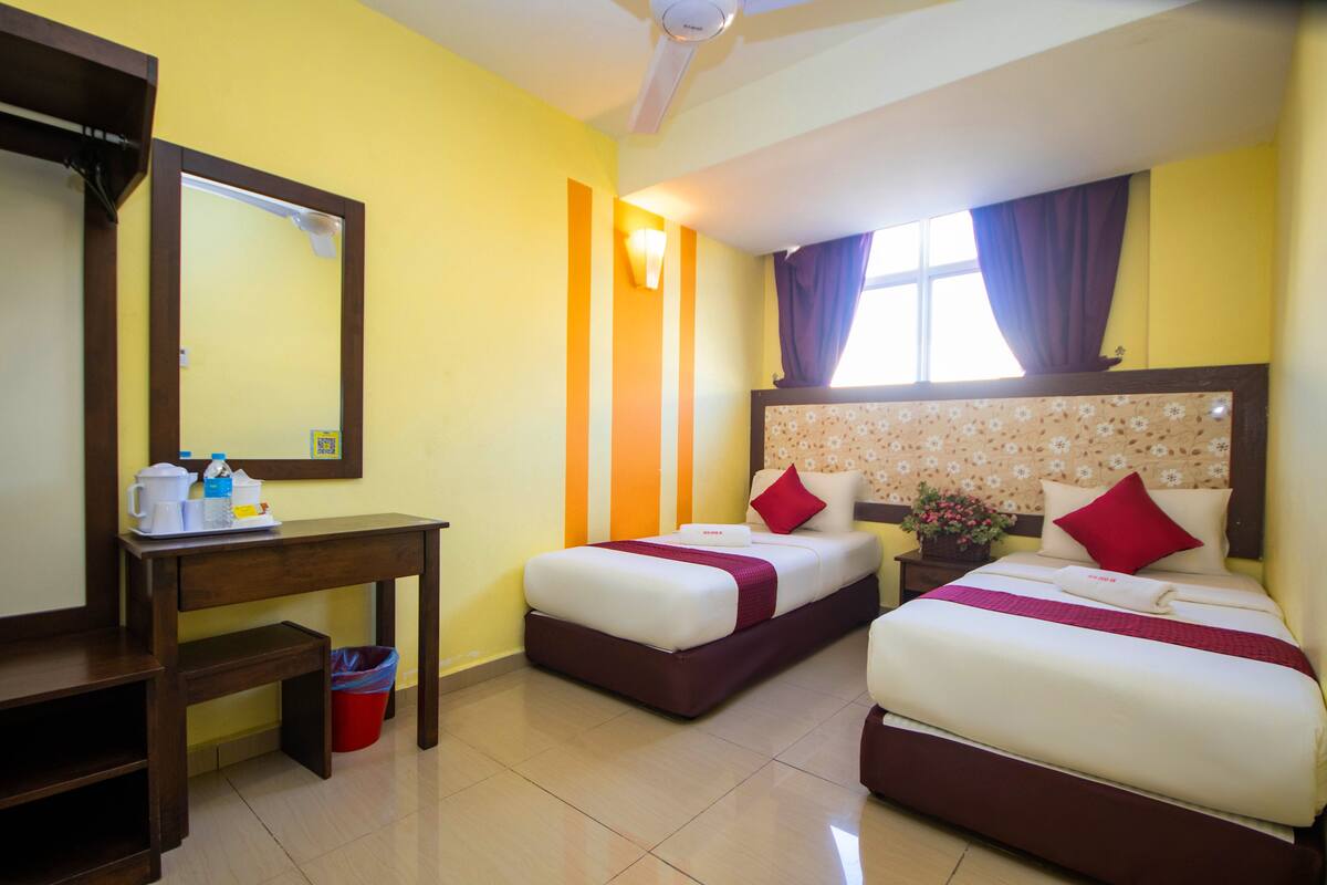Quarto de hotel com duas camas, uma escrivaninha de madeira, um cesto de lixo e uma janela com cortinas.