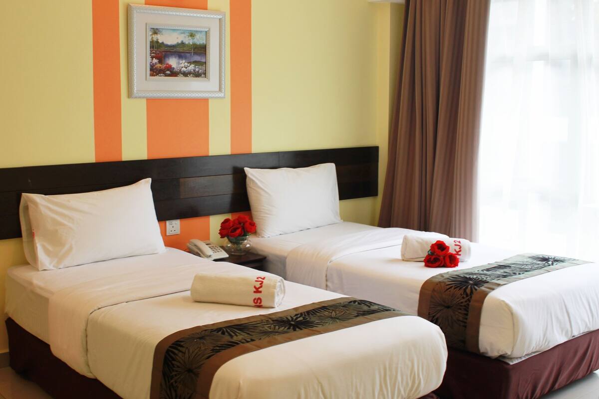 Quarto de hotel com duas camas, cada uma com roupa de cama branca e cabeceira marrom. Há uma moldura com um quadro na parede acima das camas e um vaso com flores vermelhas na mesa de cabeceira.