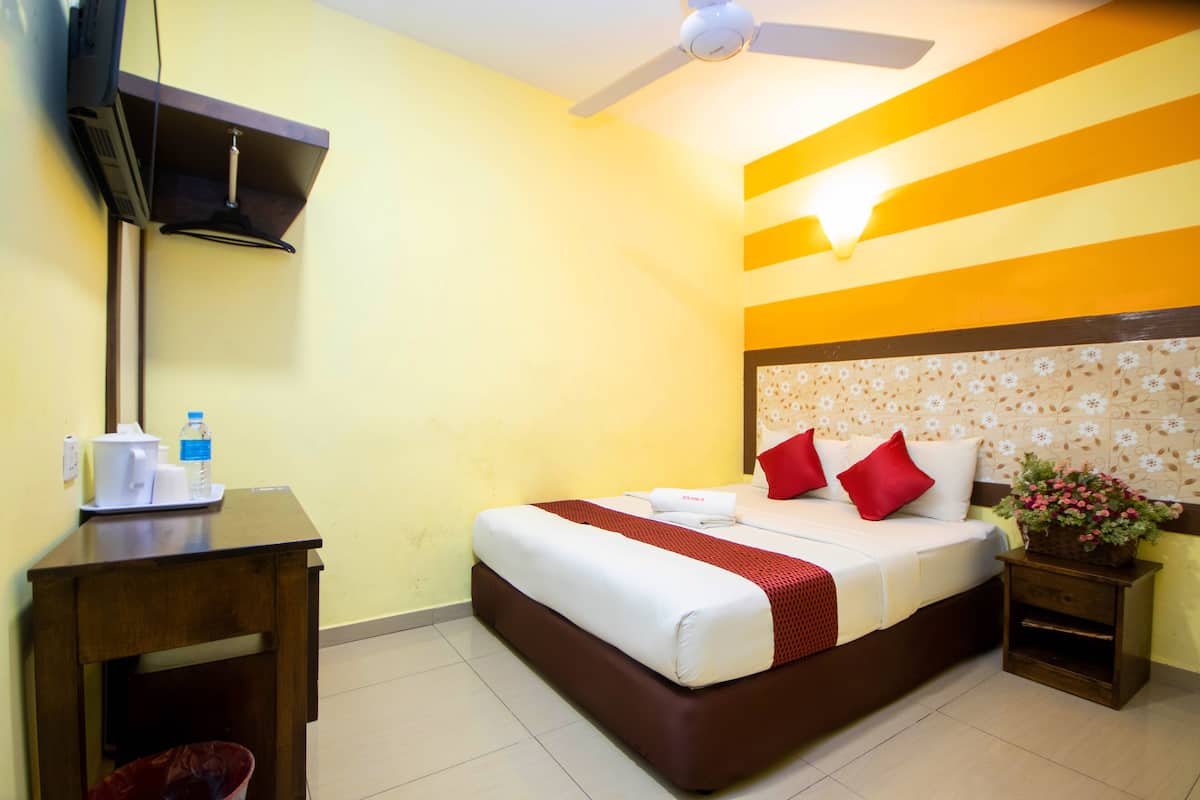 Quarto de hotel com cama, escrivaninha, televisão, ventoinha no teto e mesa de cabeceira com um vaso de flores.