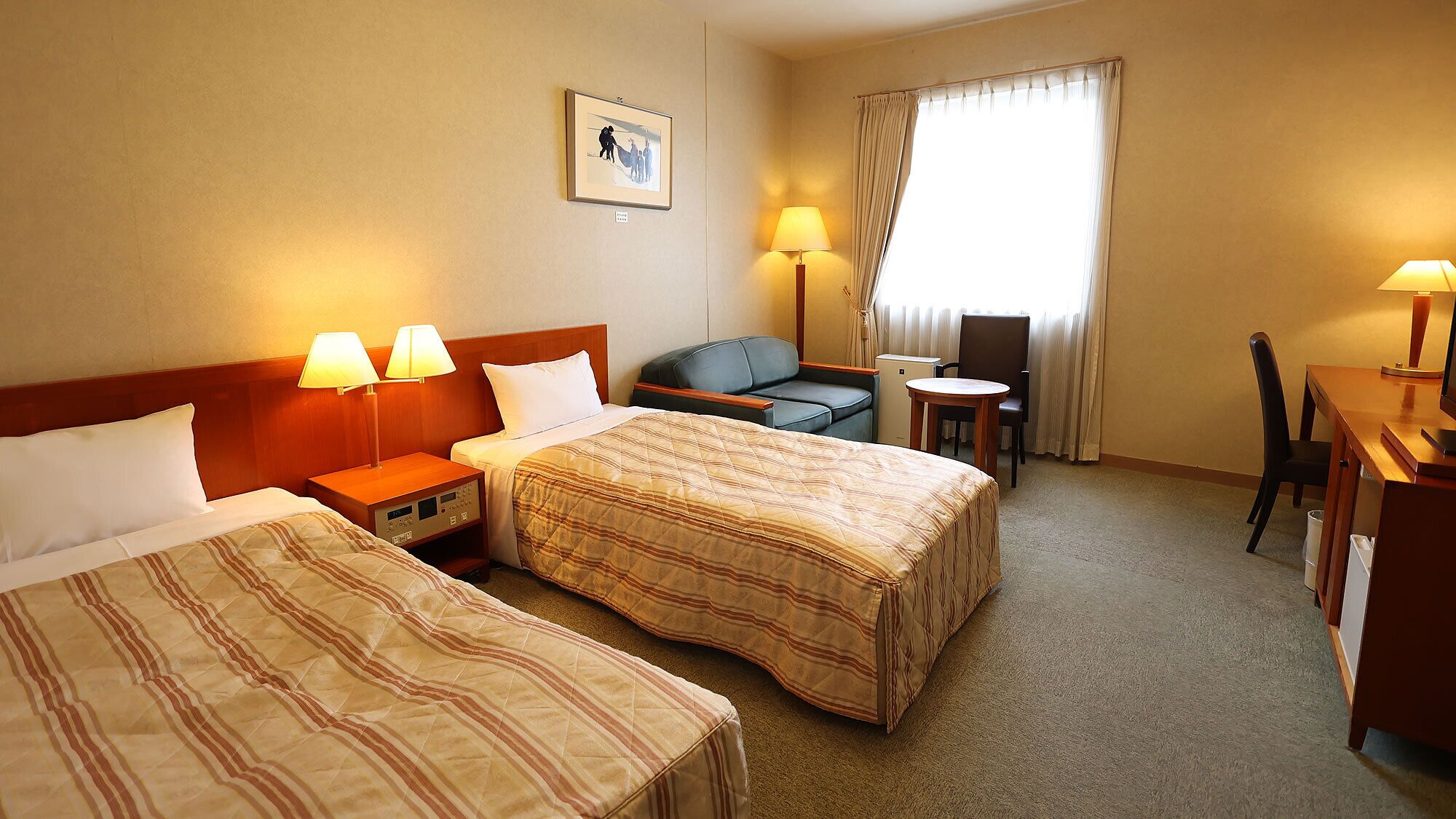 Habitación de hotel con dos camas, un escritorio, una silla, una mesita, una lámpara y una ventana con cortinas.