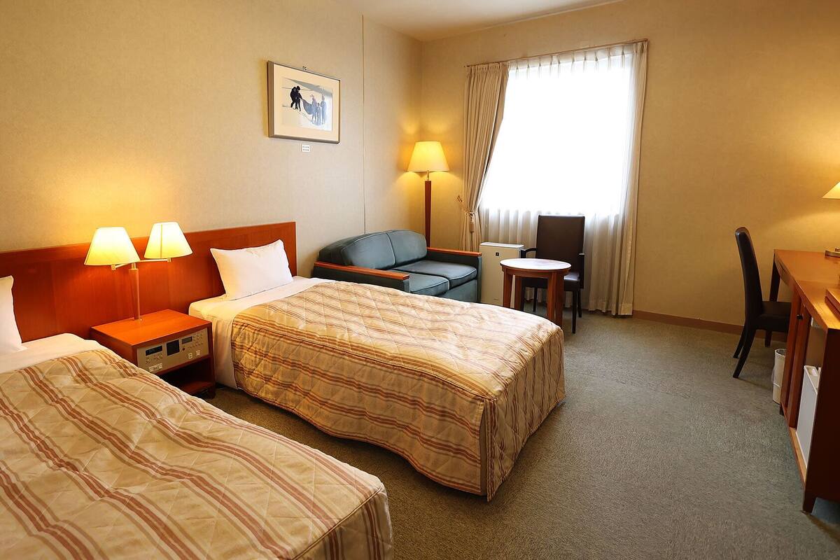 A hotel room with two beds, a desk, a chair, a small table, a lamp, and a window with curtains.