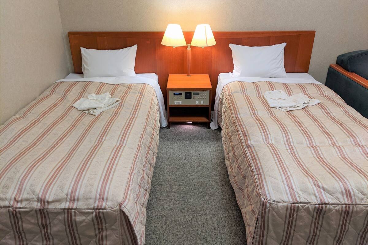 A hotel room with two single beds, each with a striped bedspread and white pillows. There is a nightstand with a lamp between the beds, and a small table with a clock.
