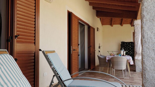 A covered outdoor seating area with a table and chairs, wooden doors, and a stone wall. - Residences Gattopardu- "Gone with the Wind" (Castiadas)