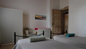 A bedroom with two beds, a wardrobe, a chair, a small table, and a painting on the wall.
