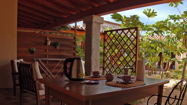 Une table en bois, sur laquelle sont posés une bouilloire, des tasses et une théière, est installée sur une terrasse d’où l’on peut admirer un jardin et un bâtiment en arrière-plan.