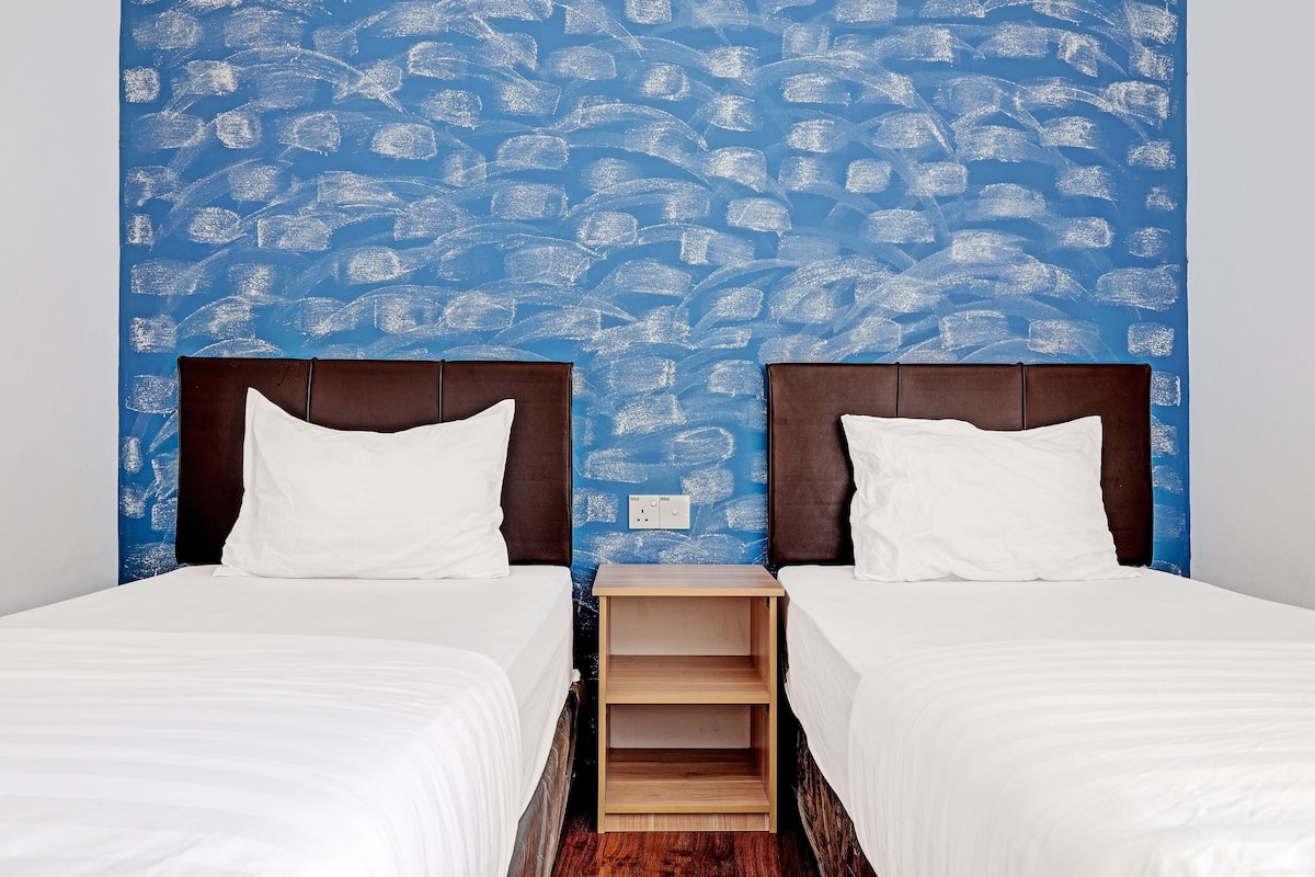 A room with two beds, each with a brown headboard and white bedding. The wall behind the beds has a blue pattern.