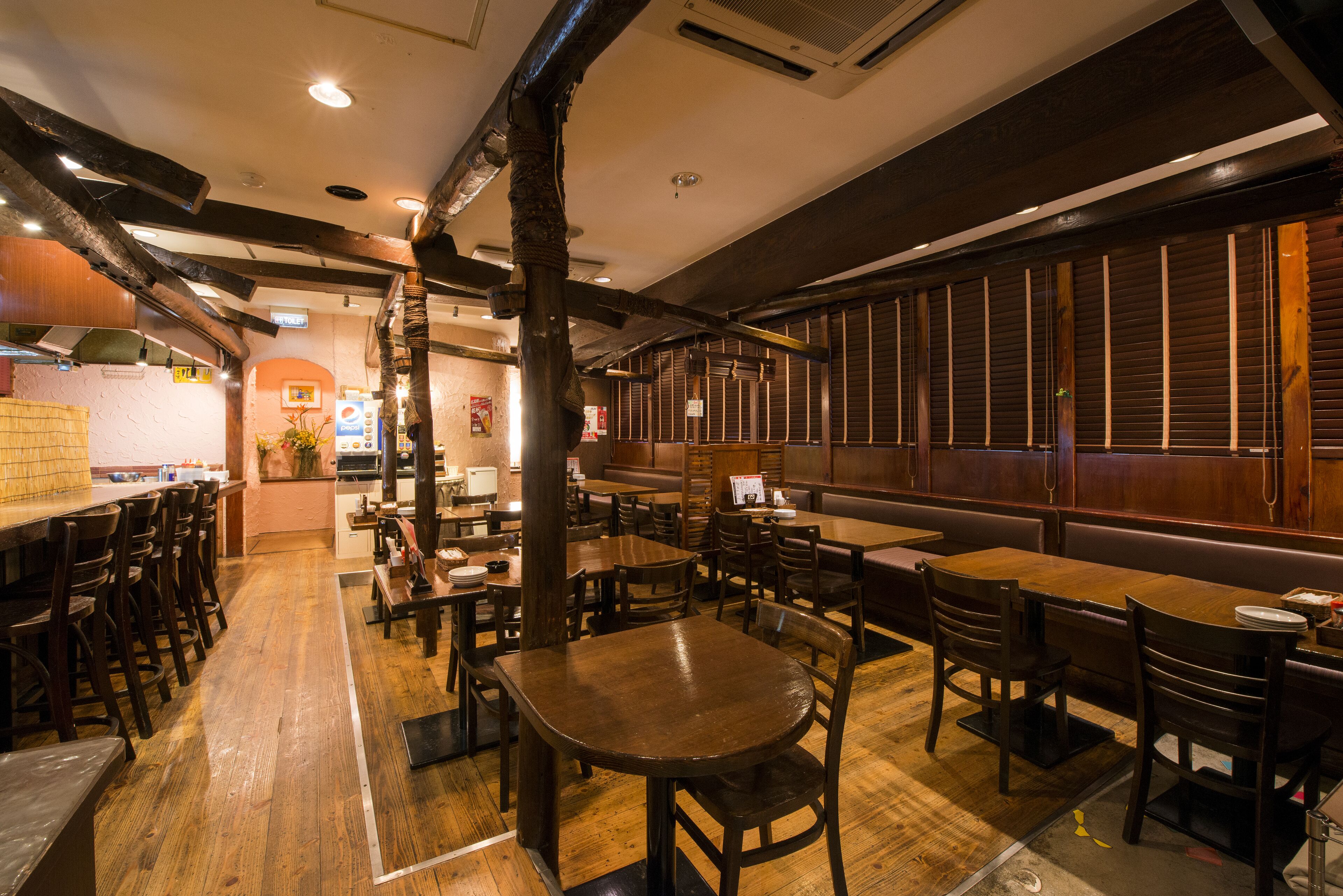 A restaurant interior with wooden tables and chairs, a kitchen area in the background, and a brick wall.