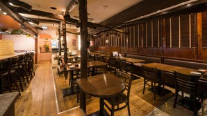 Interior de restaurante con mesas y sillas de madera, área de cocina al fondo y pared de ladrillo.