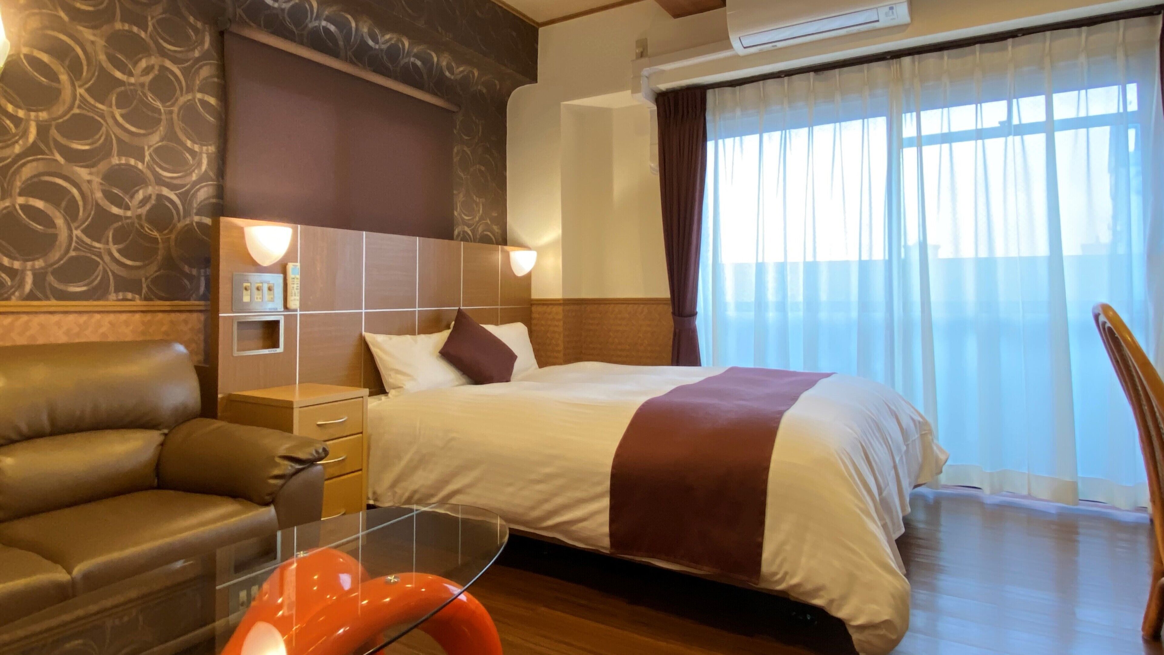 A hotel room with a bed, a desk, a chair, a nightstand, and a window with curtains.