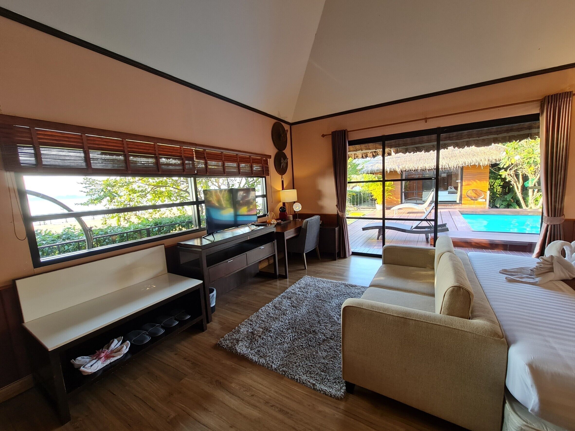 Una camera d'albergo con una scrivania in legno, una televisione, un divano e un letto. Si vede una piscina e del verde attraverso la finestra.