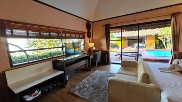 Una camera d'albergo con una scrivania in legno, una televisione, un divano e un letto. Si vede una piscina e del verde attraverso la finestra.