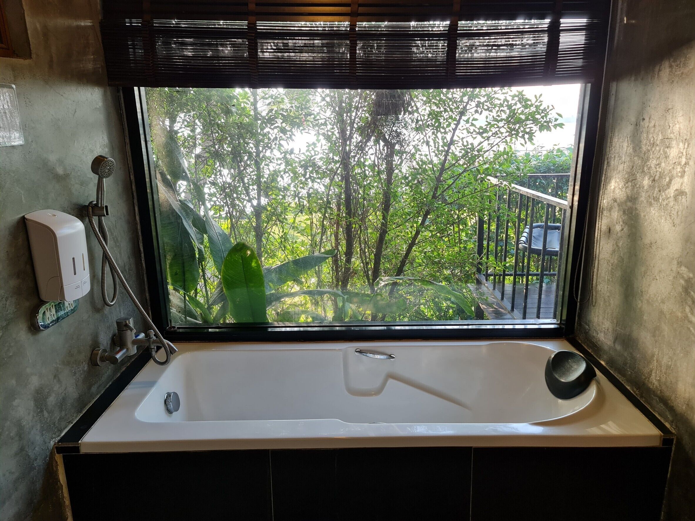 A bathroom with a large bathtub, a window with a view of trees, a hand soap dispenser, and a showerhead.