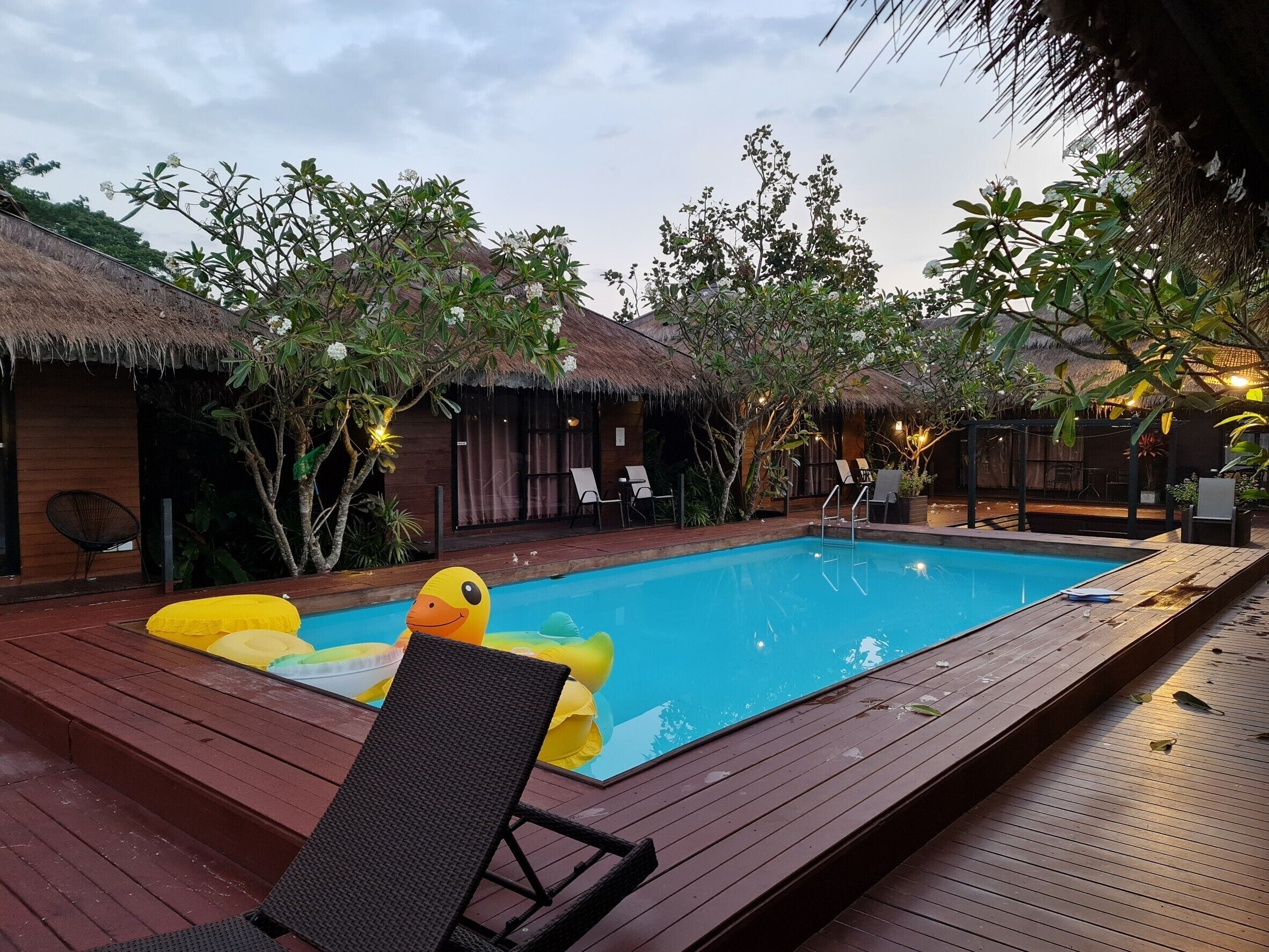 Uma área de piscina com deck de madeira, espreguiçadeiras e estruturas com telhado de palha.